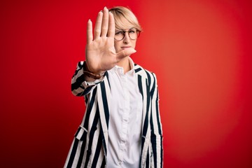 Blonde business woman with short hair wearing glasses and striped jacket over red background doing stop sing with palm of the hand. Warning expression with negative and serious gesture on the face.