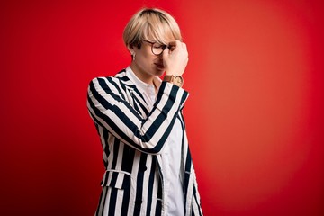 Blonde business woman with short hair wearing glasses and striped jacket over red background tired rubbing nose and eyes feeling fatigue and headache. Stress and frustration concept.