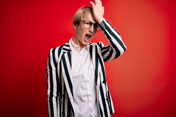 Blonde business woman with short hair wearing glasses and striped jacket over red background surprised with hand on head for mistake, remember error. Forgot, bad memory concept.