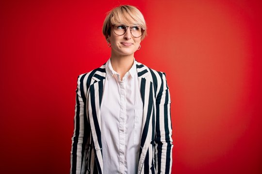 Blonde business woman with short hair wearing glasses and striped jacket over red background smiling looking to the side and staring away thinking.