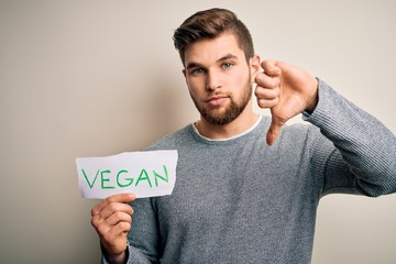 Young blond veggie man with beard and blue eyes holding paper with vegan message with angry face,...