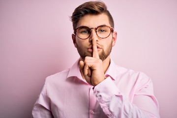 Young handsome blond man with beard and blue eyes wearing pink shirt and glasses asking to be quiet with finger on lips. Silence and secret concept.