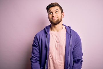 Fototapeta premium Young blond man with beard and blue eyes wearing purple sweatshirt over pink background smiling looking to the side and staring away thinking.