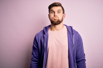 Young blond man with beard and blue eyes wearing purple sweatshirt over pink background puffing cheeks with funny face. Mouth inflated with air, crazy expression.