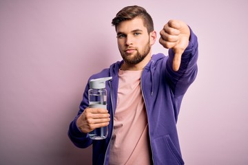 Young blond sporty man with beard and blue eyes doing sport holding bottle of water with angry...