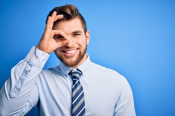 Young blond businessman with beard and blue eyes wearing elegant shirt and tie standing doing ok gesture with hand smiling, eye looking through fingers with happy face.