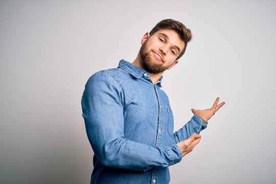 Young handsome blond man with beard and blue eyes wearing casual denim shirt Inviting to enter smiling natural with open hand