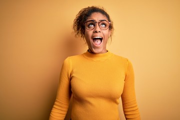 Young beautiful african american girl wearing sweater and glasses over yellow background angry and mad screaming frustrated and furious, shouting with anger. Rage and aggressive concept.