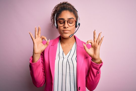 Young african american call center agent girl wearing glasses working using headset relax and smiling with eyes closed doing meditation gesture with fingers. Yoga concept.