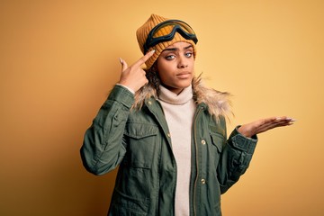 Young african american afro skier girl wearing snow sportswear and ski goggles confused and annoyed with open palm showing copy space and pointing finger to forehead. Think about it.