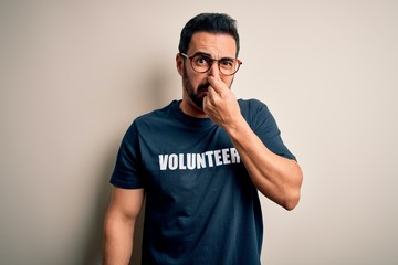 Handsome man with beard wearing t-shirt with volunteer message over white background smelling something stinky and disgusting, intolerable smell, holding breath with fingers on nose. Bad smell