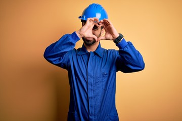 Mechanic man with beard wearing blue uniform and safety helmet over yellow background Doing heart shape with hand and fingers smiling looking through sign