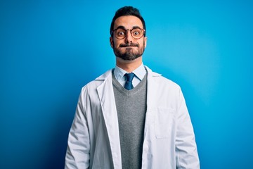 Young handsome doctor man with beard wearing coat and glasses over blue background puffing cheeks with funny face. Mouth inflated with air, crazy expression.