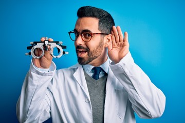 Young handsome optical man with beard holding optometry glasses over blue background smiling with hand over ear listening an hearing to rumor or gossip. Deafness concept.