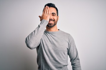 Young handsome man with beard wearing casual sweater standing over white background covering one eye with hand, confident smile on face and surprise emotion.