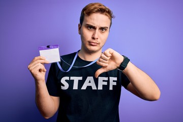 Young handsome redhead worker man wearing staff t-shirt uniform holding id card pass with angry face, negative sign showing dislike with thumbs down, rejection concept