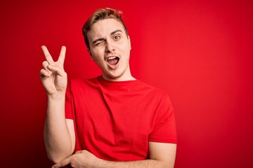 Naklejka premium Young handsome redhead man wearing casual t-shirt over isolated red background smiling with happy face winking at the camera doing victory sign. Number two.