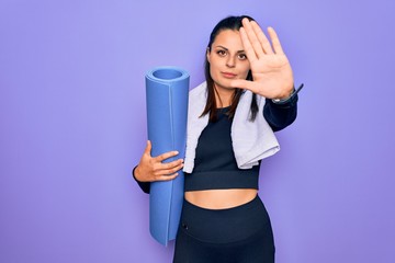 Young beautiful brunette sporty woman using sport towel hoding mat to do yoga with open hand doing stop sign with serious and confident expression, defense gesture