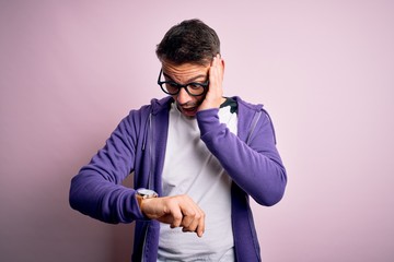 Young handsome man wearing purple sweatshirt and glasses standing over pink background Looking at the watch time worried, afraid of getting late