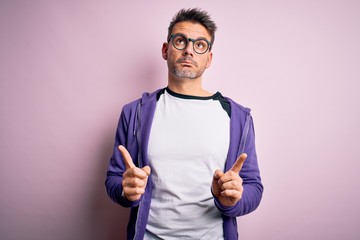 Young handsome man wearing purple sweatshirt and glasses standing over pink background Pointing up looking sad and upset, indicating direction with fingers, unhappy and depressed.
