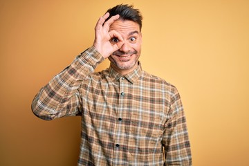 Obraz premium Young handsome man wearing casual shirt standing over isolated yellow background doing ok gesture with hand smiling, eye looking through fingers with happy face.