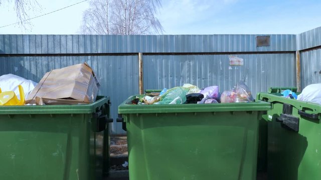 Large green containers full of rubbish in plastic bags located near metal barrier on sunny day on street. The concept of ecology and environmental protection.