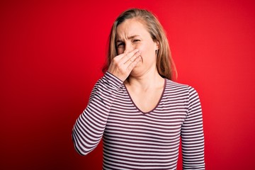 Young beautiful blonde woman wearing casual striped t-shirt over isolated red background smelling...