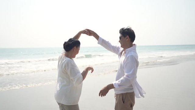 HD Slow motion of happy senior Asian couple man and woman holding hands and dancing together on the beach with smiling. Retired old age family relaxing in romantic marriage anniversary travel.