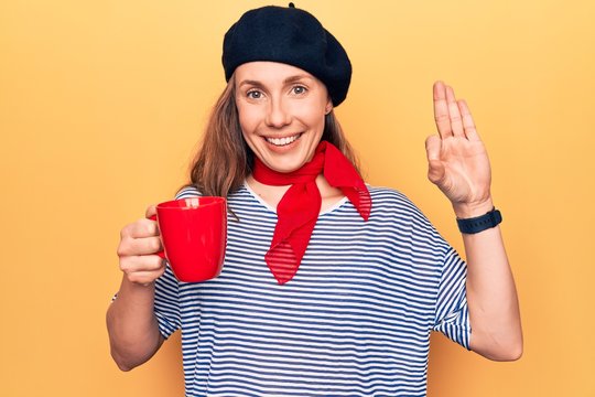 Young beautiful blonde woman wearing fashion beret drinking a cup of coffee doing ok sign with fingers, smiling friendly gesturing excellent symbol