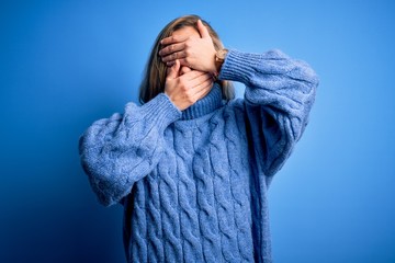 Young beautiful blonde woman wearing casual turtleneck sweater over blue background Covering eyes and mouth with hands, surprised and shocked. Hiding emotion