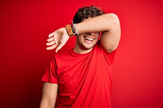 Young blond handsome man with curly hair wearing casual t-shirt over red background covering eyes with arm smiling cheerful and funny. Blind concept.