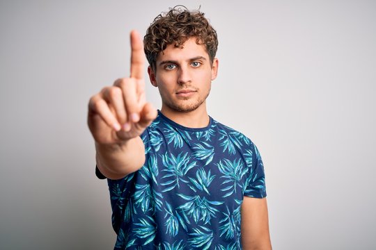 Young blond handsome man on vacation with curly hair wearing casual summer t-shirt Pointing with finger up and angry expression, showing no gesture