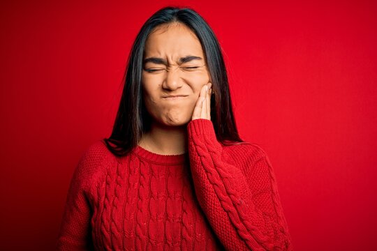 Young beautiful asian woman wearing casual sweater standing over isolated red background touching mouth with hand with painful expression because of toothache or dental illness on teeth. Dentist