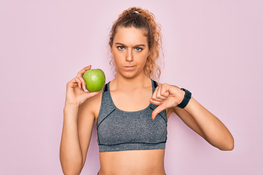 Young beautiful blonde sporty woman with blue eyes holding healthy green apple fruit with angry face, negative sign showing dislike with thumbs down, rejection concept