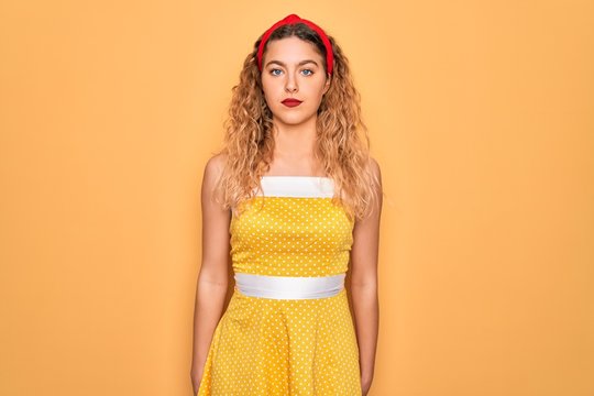 Beautiful blonde pin-up woman with blue eyes wearing diadem standing over yellow background Relaxed with serious expression on face. Simple and natural looking at the camera.