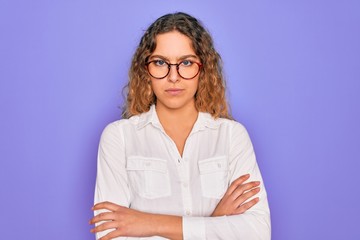 Young beautiful woman with blue eyes wearing casual shirt and glasses over purple background skeptic and nervous, disapproving expression on face with crossed arms. Negative person.