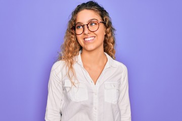 Obraz premium Young beautiful woman with blue eyes wearing casual shirt and glasses over purple background looking away to side with smile on face, natural expression. Laughing confident.