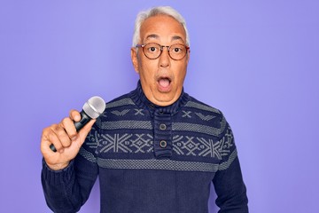 Middle age senior grey-haired singer man singing using music microphone over purple background scared in shock with a surprise face, afraid and excited with fear expression