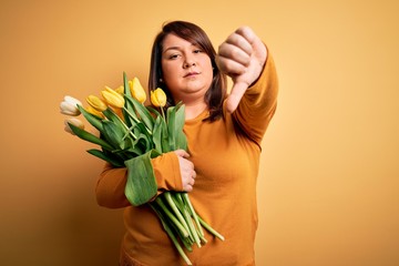 Beautiful plus size woman holding romantic bouquet of natural tulips flowers over yellow background looking unhappy and angry showing rejection and negative with thumbs down gesture. Bad expression.