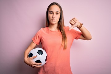 Young beautiful player woman playing soccer holding football ball  over pink background with angry face, negative sign showing dislike with thumbs down, rejection concept