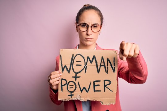 Young beautiful activist woman asking for women rights holding banner with power message pointing with finger to the camera and to you, hand sign, positive and confident gesture from the front
