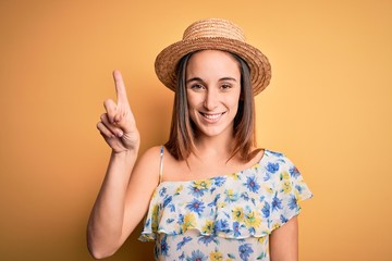 Young beautiful woman wearing casual t-shirt and summer hat over isolated yellow background showing and pointing up with finger number one while smiling confident and happy.