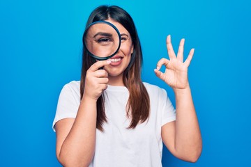 Young beautiful brunette woman using magnifying glass over isolated blue background doing ok sign with fingers, smiling friendly gesturing excellent symbol
