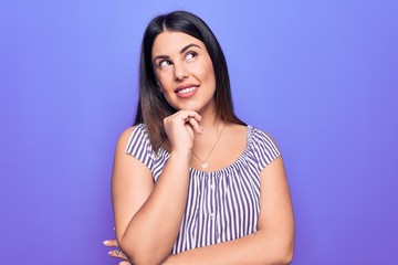Fototapeta premium Young beautiful brunette woman wearing casual striped t-shirt standing over purple background smiling looking confident at the camera with crossed arms and hand on chin. Thinking positive.
