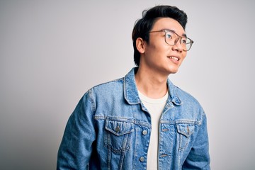 Young handsome chinese man wearing denim jacket and glasses over white background looking away to side with smile on face, natural expression. Laughing confident.
