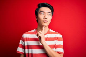 Young handsome chinese man wearing casual striped t-shirt standing over red background Thinking concentrated about doubt with finger on chin and looking up wondering