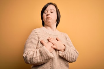 Young down syndrome woman wearing casual sweater over yellow background smiling with hands on chest with closed eyes and grateful gesture on face. Health concept.