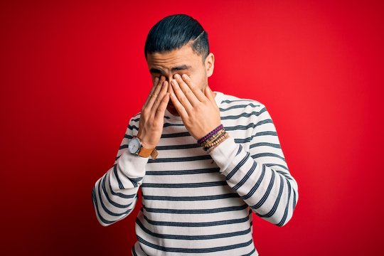 Young brazilian man wearing casual striped t-shirt standing over isolated red background rubbing eyes for fatigue and headache, sleepy and tired expression. Vision problem