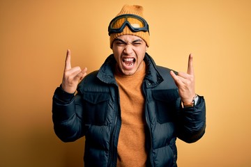 Young brazilian skier man wearing snow sportswear and ski goggles over yellow background shouting...