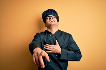 Young brazilian chef man wearing cooker uniform and hat over isolated yellow background laughing at you, pointing finger to the camera with hand over body, shame expression